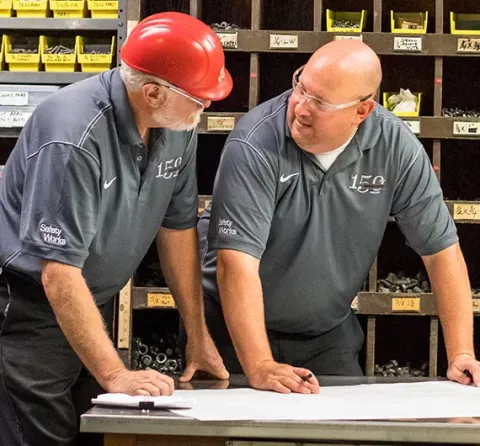 Two employees discuss a project blueprints at a workstation.